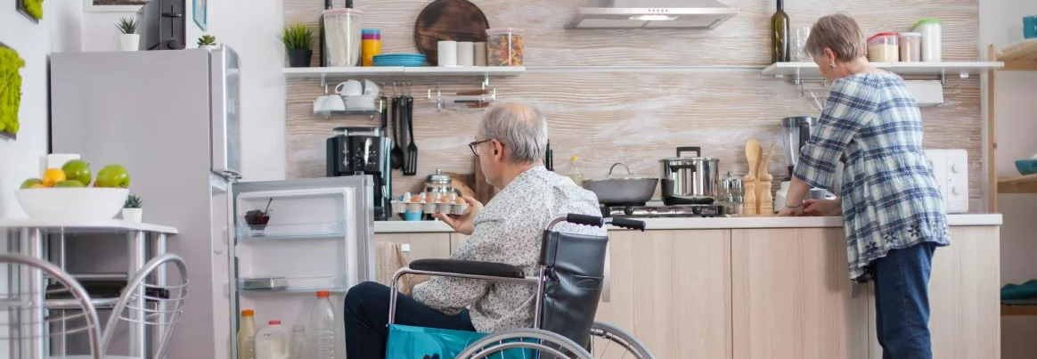 Mann im Rollstuhl holt Eierkarton aus dem Kühlschrank für seine Frau in der Küche.